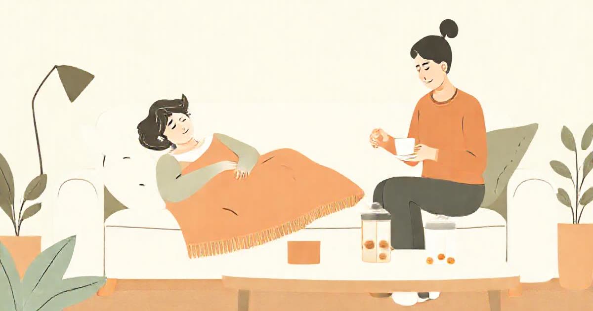 A person resting on a sofa with a blanket while their partner prepares tea, pill organiser on the table.