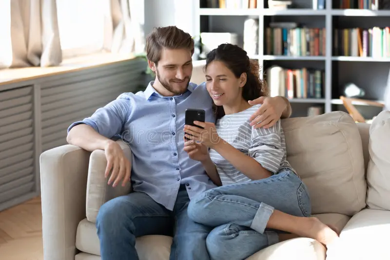 Happy young Indian couple sitting closely on sofa smiling warmly at smartphone receiving gentle helpful reminder from EvenUS app, cozy living room setting, positive relationship harmony